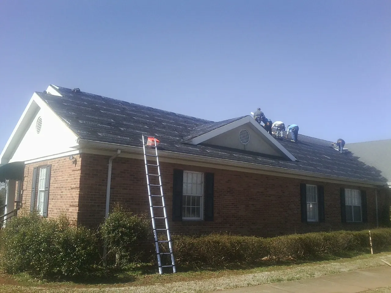 Crew tearing shingles off a brick ranch roof with a ladder set — Emergency Roof Repair work by our crew in Weedsport