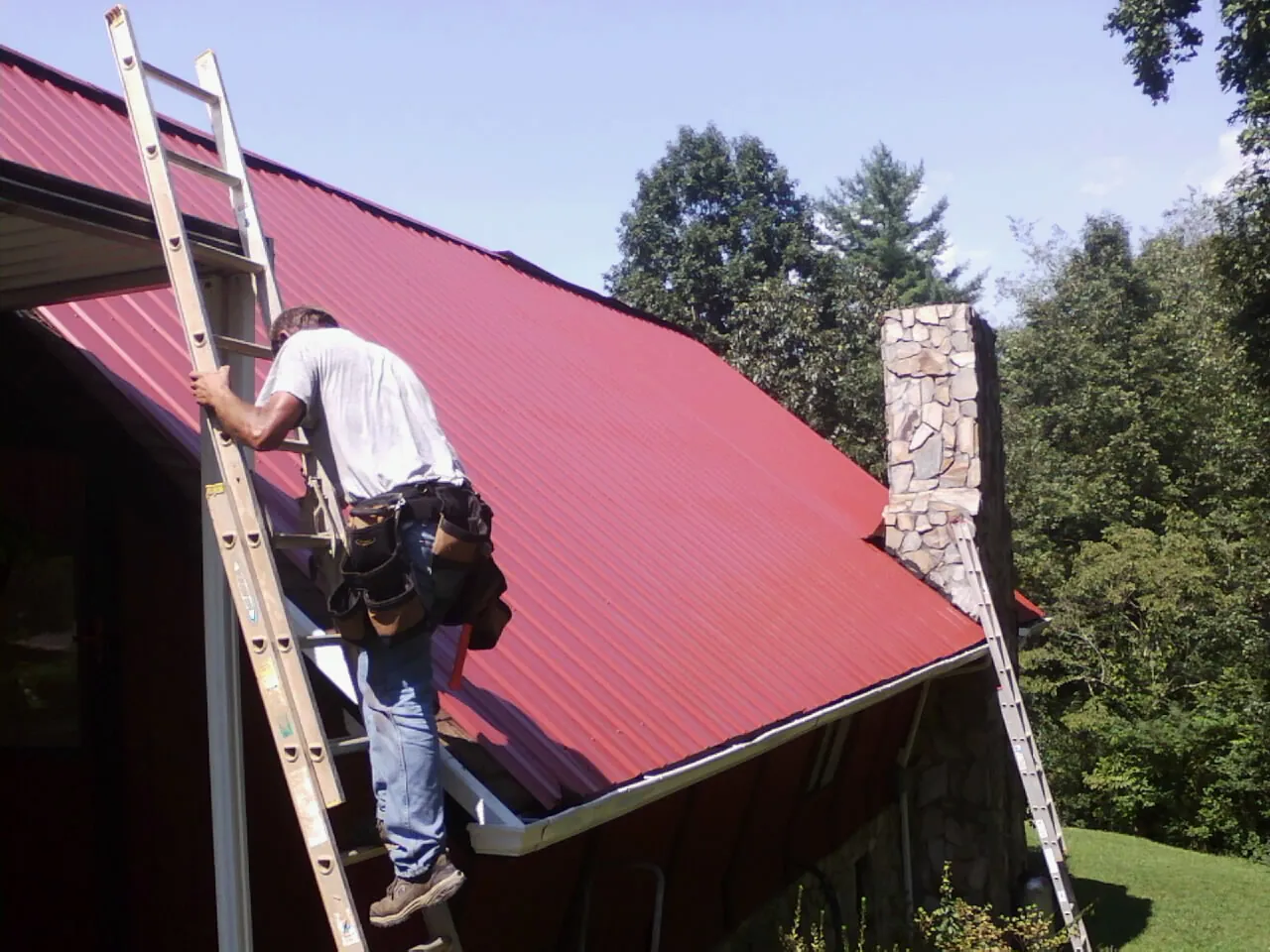 Roofer on a ladder beside a red metal roof and stone chimney — Soffit & Fascia Repair by Roofing Brothers in Weedsport